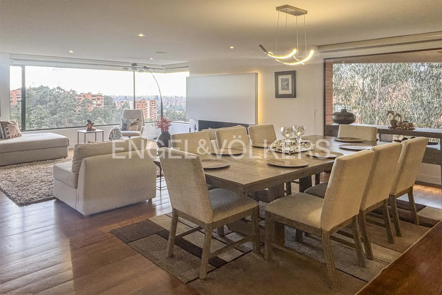 Open-concept living and dining area with a large window view. A wooden table with beige chairs sits under a modern light fixture.