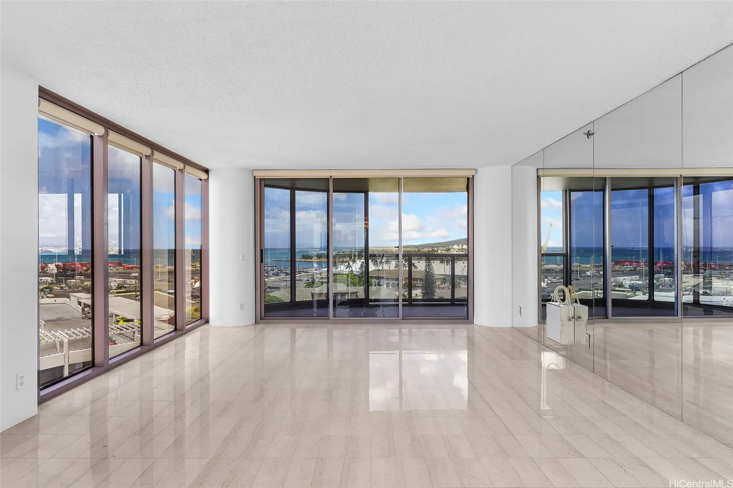Empty room with light tile floors, floor-to-ceiling windows, and a mirrored wall overlooking a harbor and ocean view.