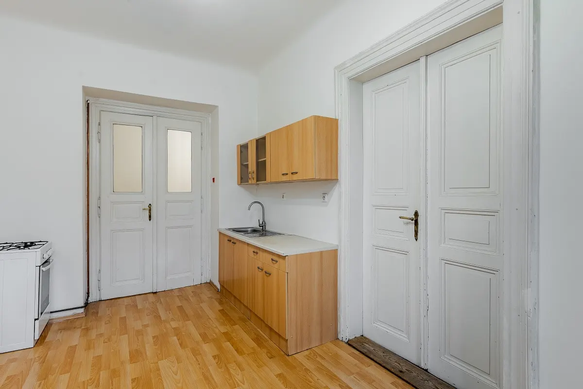 A bright kitchen with wood cabinets, a sink, and a white stove. Two sets of white doors are visible. The floor is wood.