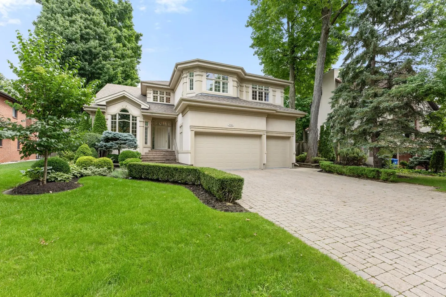 Two-story beige house with a two-car garage, brick driveway, green lawn, and manicured bushes.