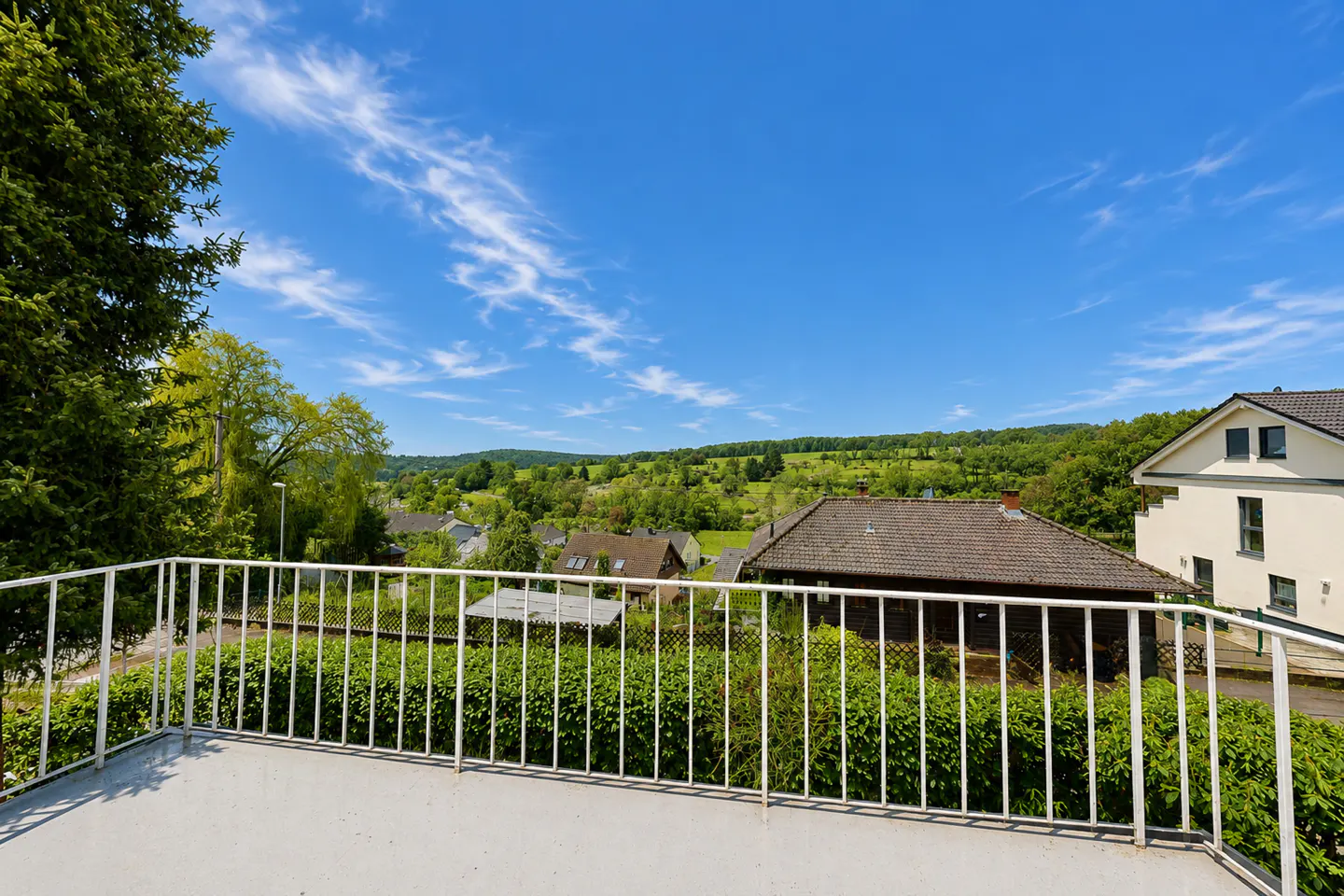View from a balcony with white railings overlooking green hills, houses, and a blue sky with wispy clouds.