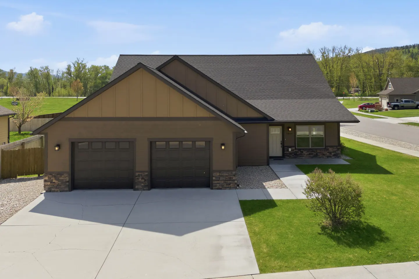 A single-story brown house with a two-car garage, a gray roof, and a green lawn on a sunny day.