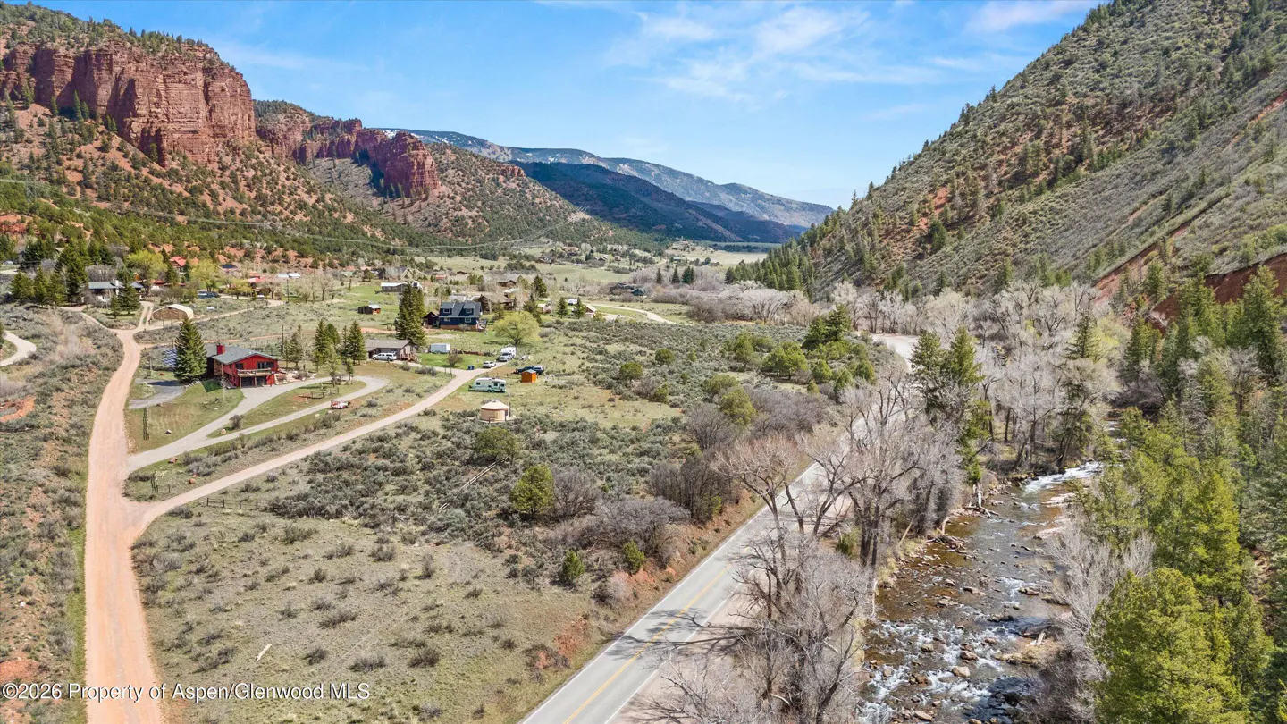 Aerial view of a valley with red rock cliffs, homes, a road, and a river under a blue sky.