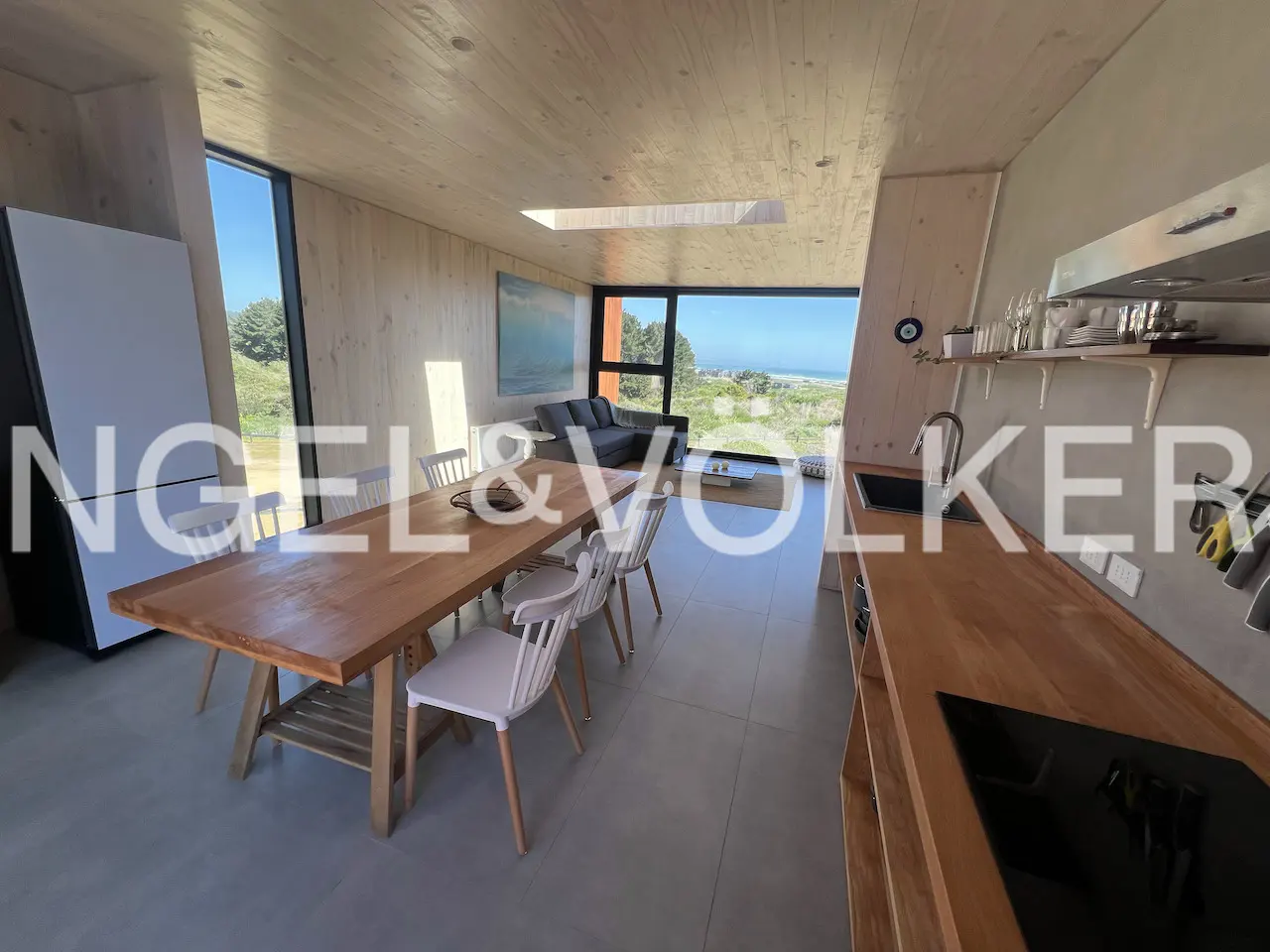Open-concept living space with wood walls, a dining table with white chairs, and a kitchen with wood countertops. Ocean view through large windows.