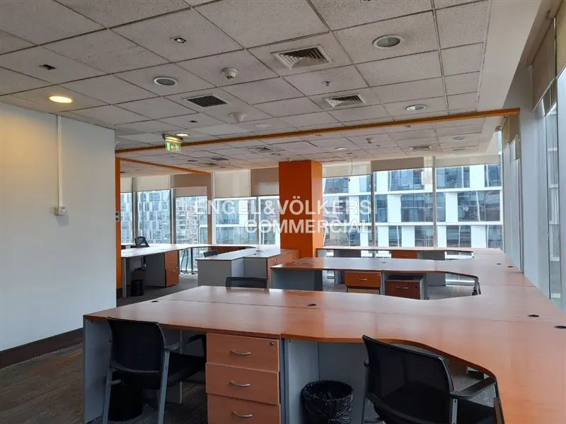 Bright office space with desks, chairs, and large windows. Orange accents on desks and pillars. Engel & Völkers Commercial logo visible.
