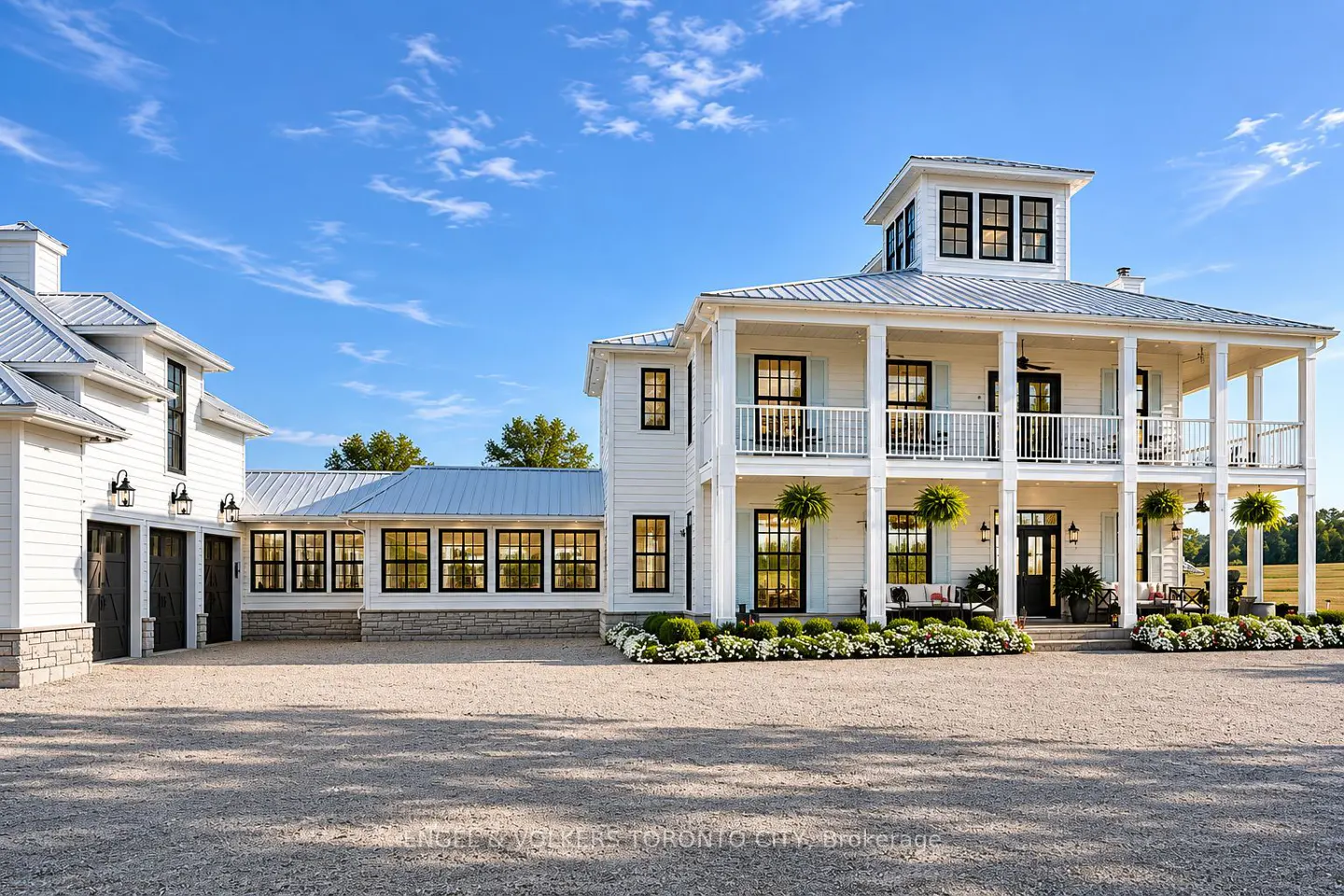 Two-story white farmhouse with a metal roof, black window frames, and a gravel driveway under a blue sky.