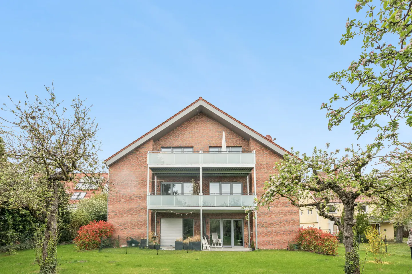 Two-story brick house with two balconies, green lawn, and trees with white blossoms under a blue sky.