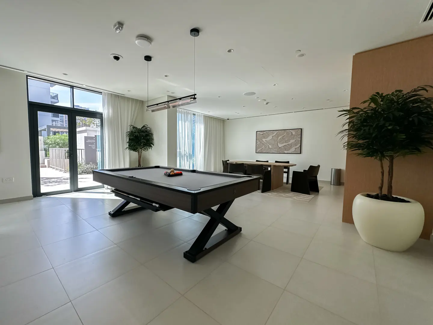 Bright, modern recreation room with a pool table, dining table, and potted plants. Light tile flooring and large windows.