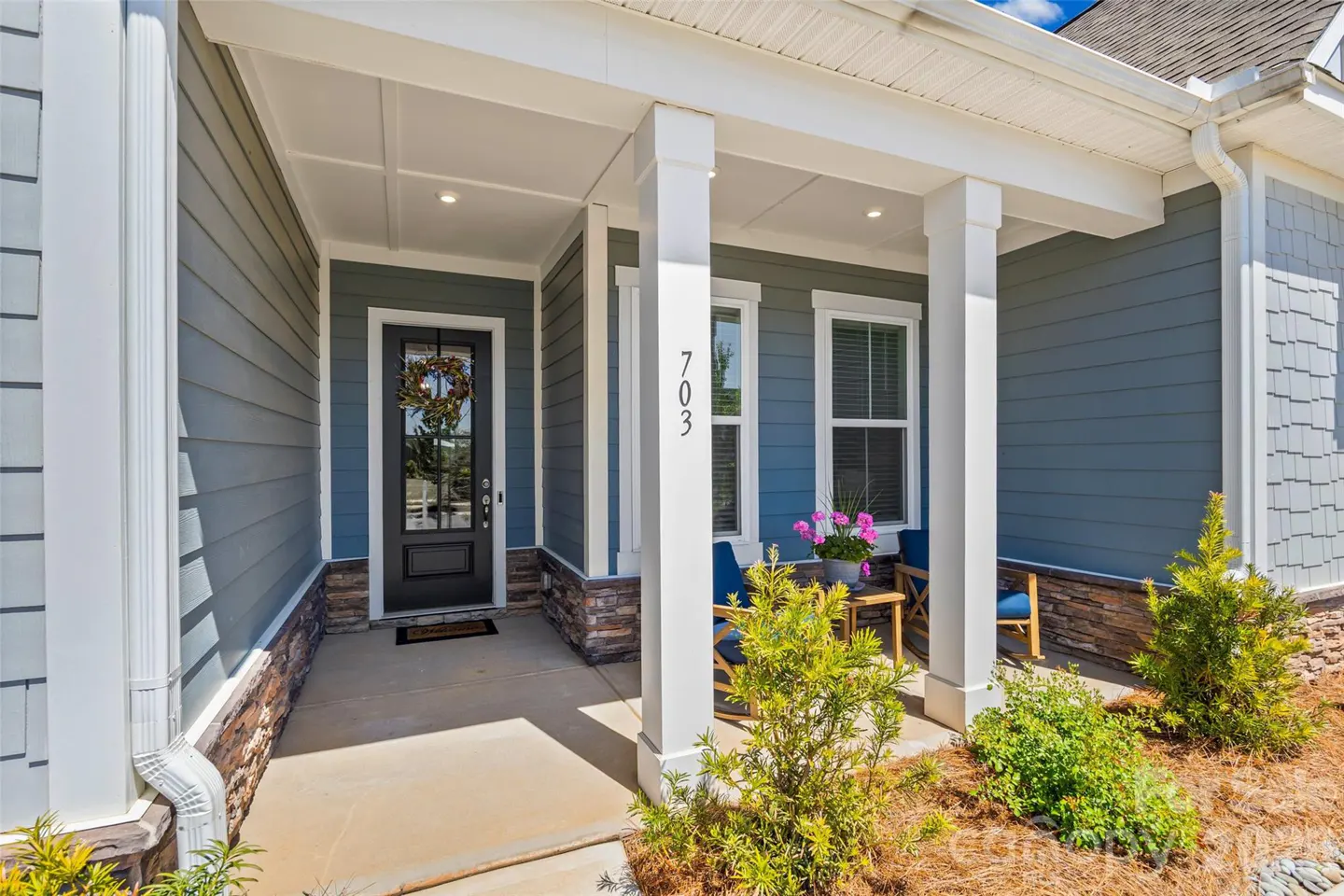 Covered front porch of a blue-sided home with stone accents, a dark front door with a wreath, and two blue patio chairs.