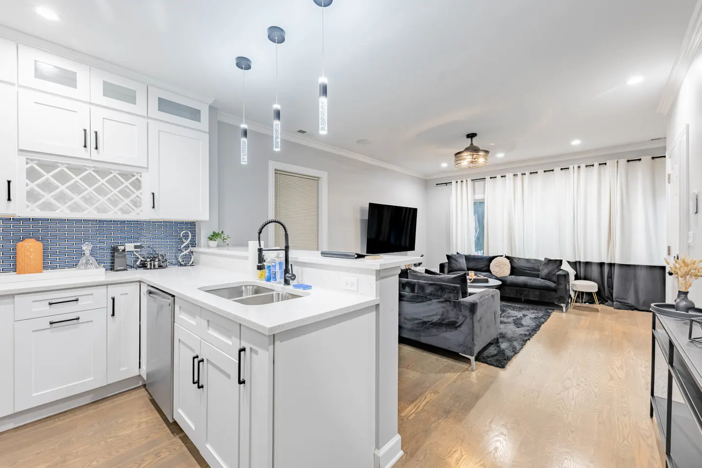 Open concept living space with white kitchen cabinets, blue backsplash, and gray velvet sofas.