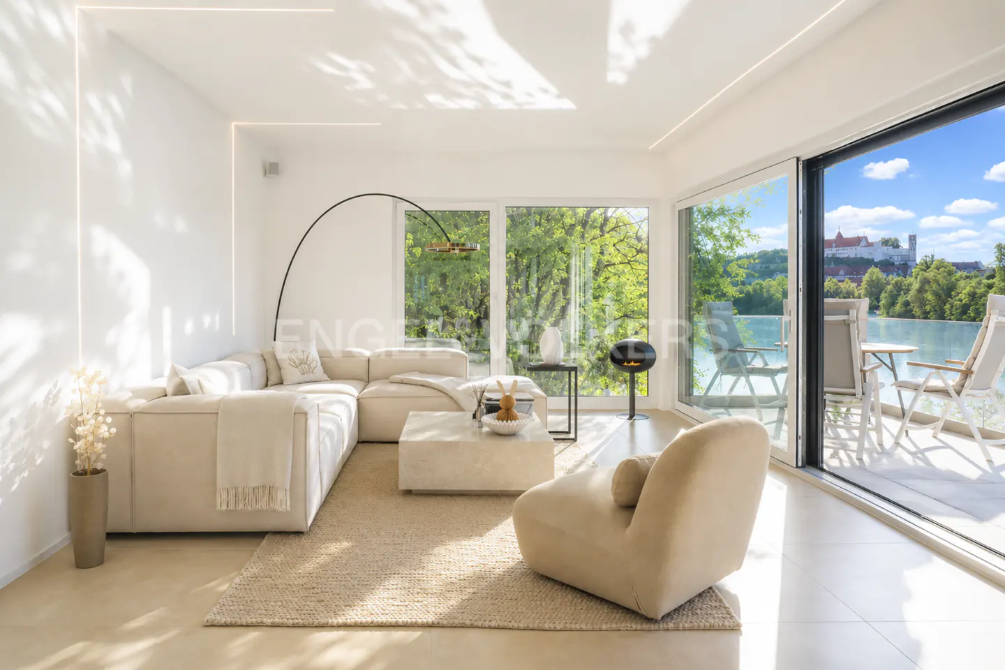 Modern living room with beige sectional, armchair, and large windows overlooking a lake and castle under a blue sky.