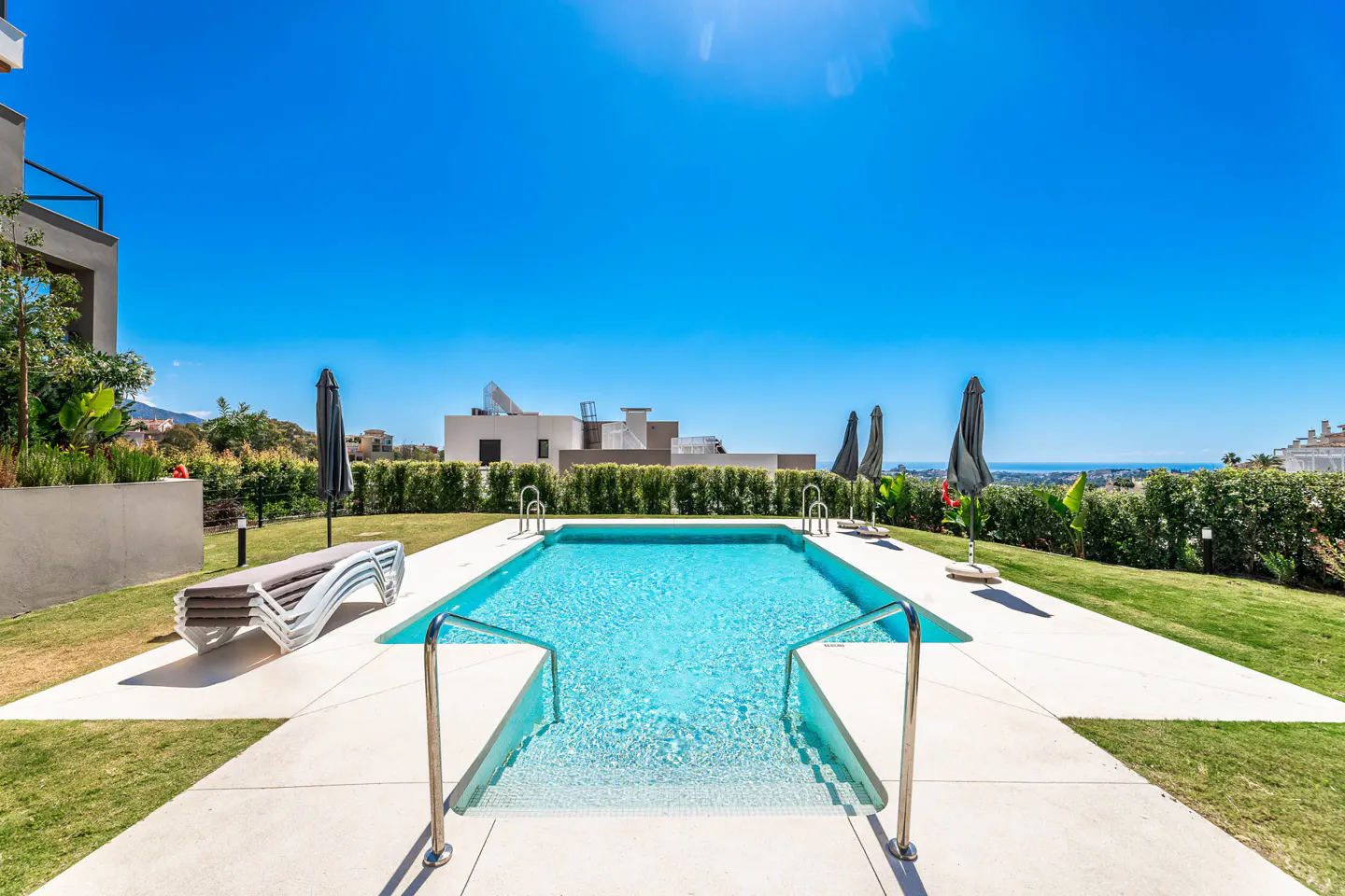 Outdoor pool with turquoise water, metal handrails, and white tile surround. Lounge chairs and umbrellas sit on the green lawn under a clear blue sky.