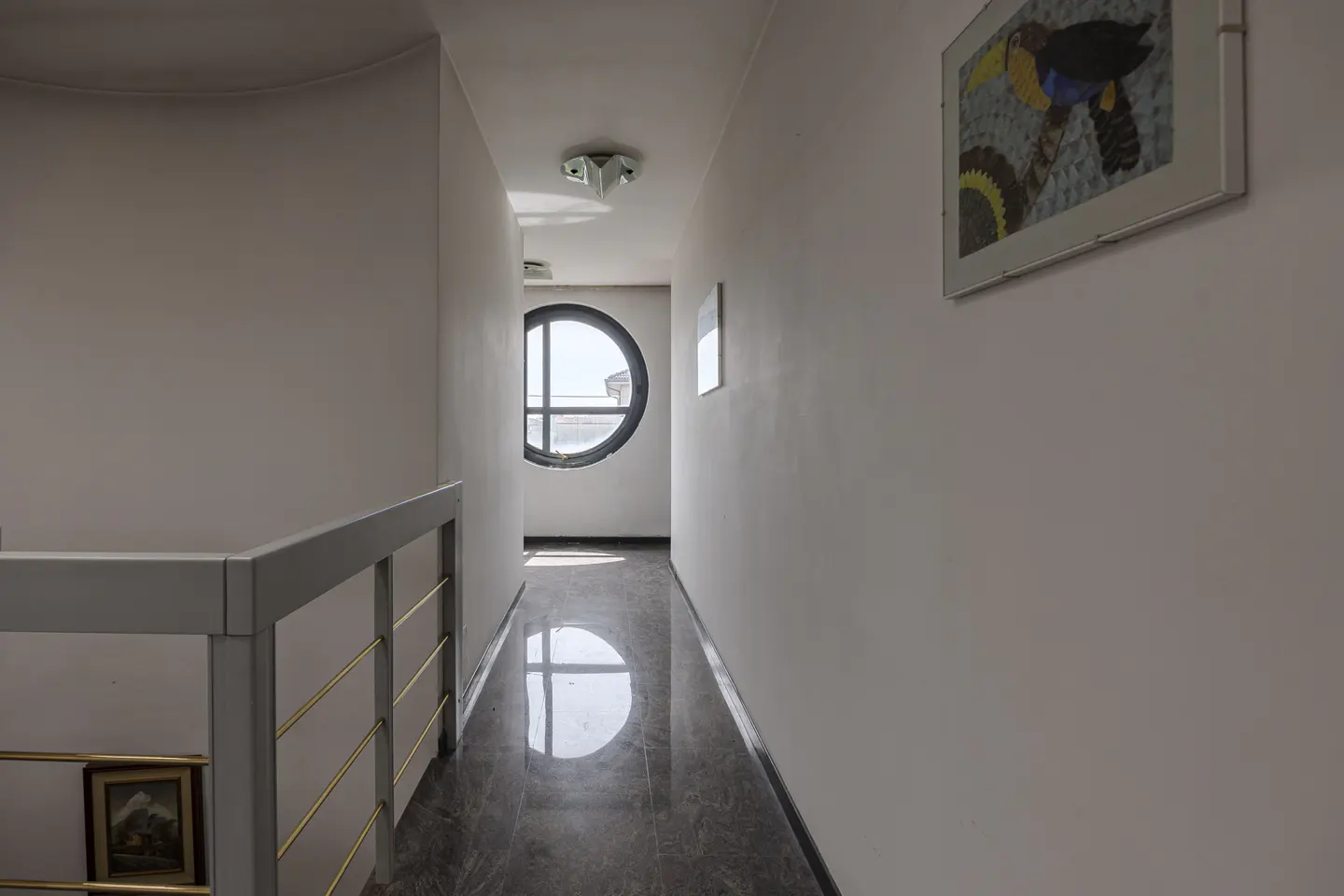 A bright hallway with white walls, a dark marble floor, a round window at the end, and a modern railing on the left.