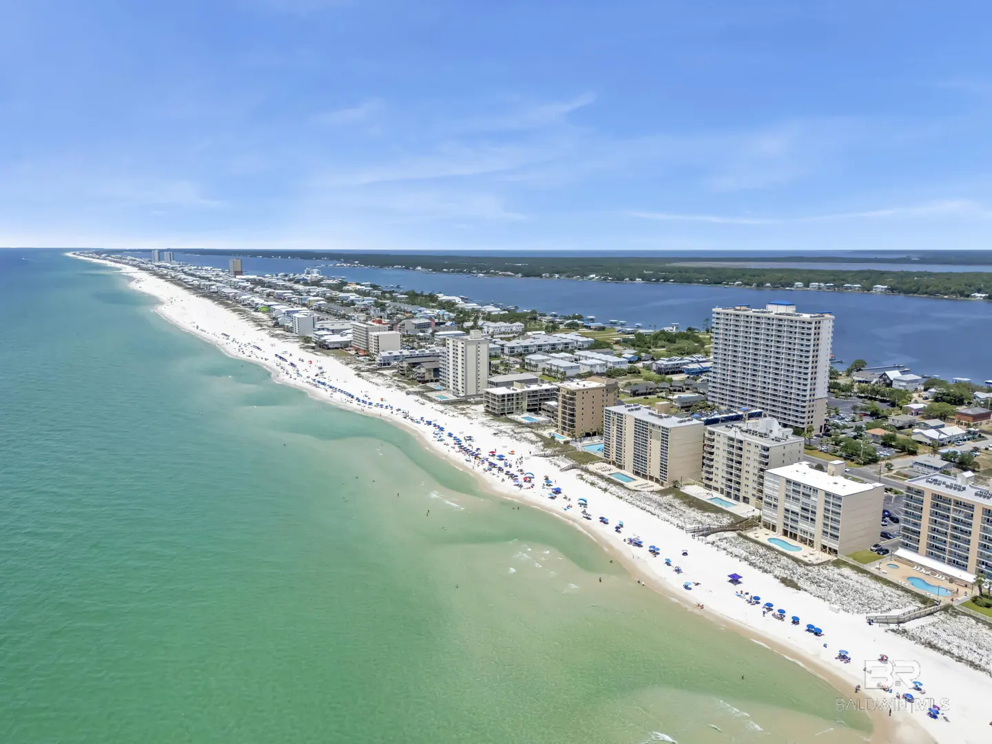 Aerial view of a white sand beach with turquoise water, lined with condos and buildings under a blue sky.