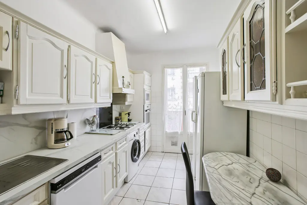 Bright, narrow kitchen with white cabinets, appliances, and marble countertops. A window is visible at the end of the room.
