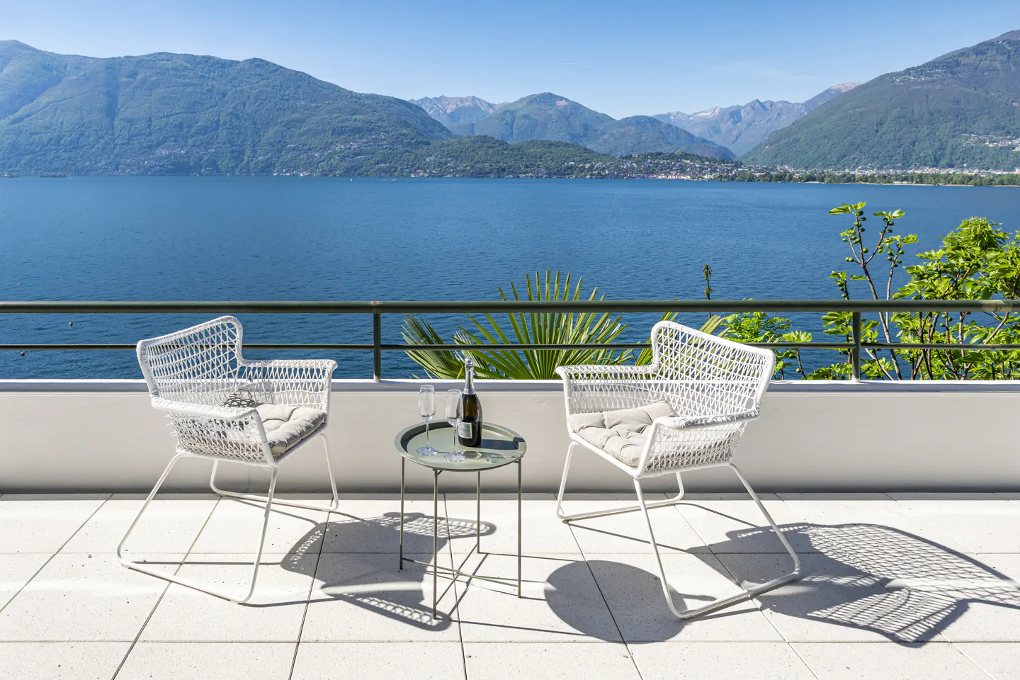 Balcony with two white chairs, a table with champagne and glasses, overlooking a blue lake and mountains on a sunny day.