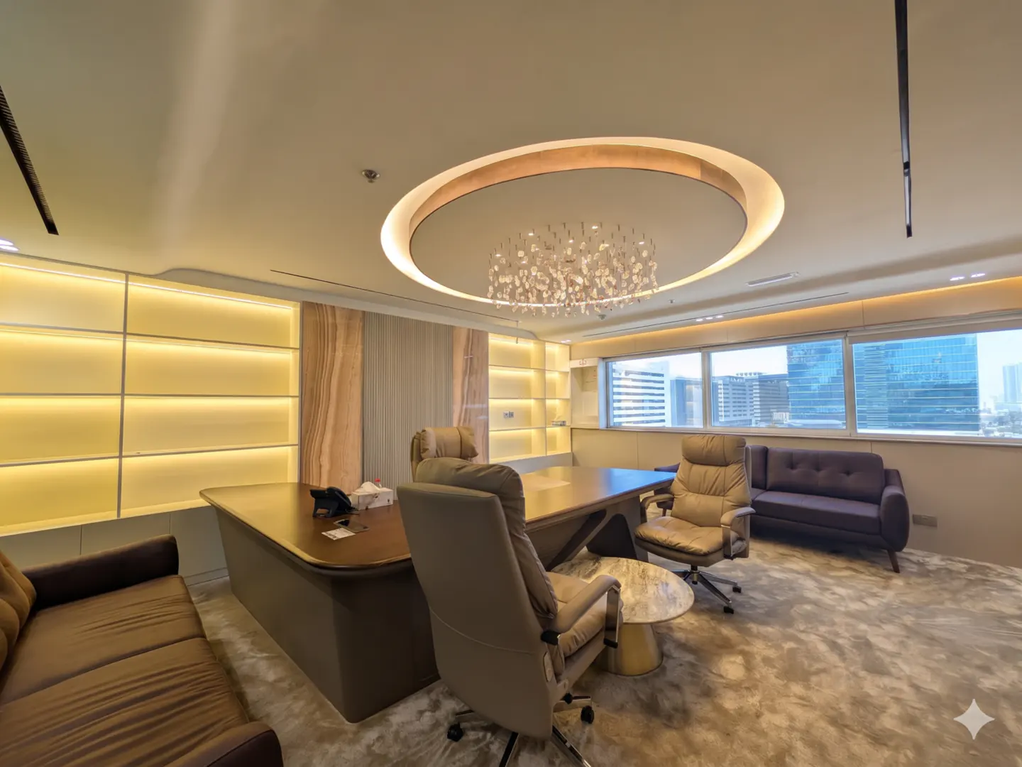 A modern office space with a large desk, leather chairs, and a brown sofa. A chandelier hangs from the ceiling. Windows offer a city view.