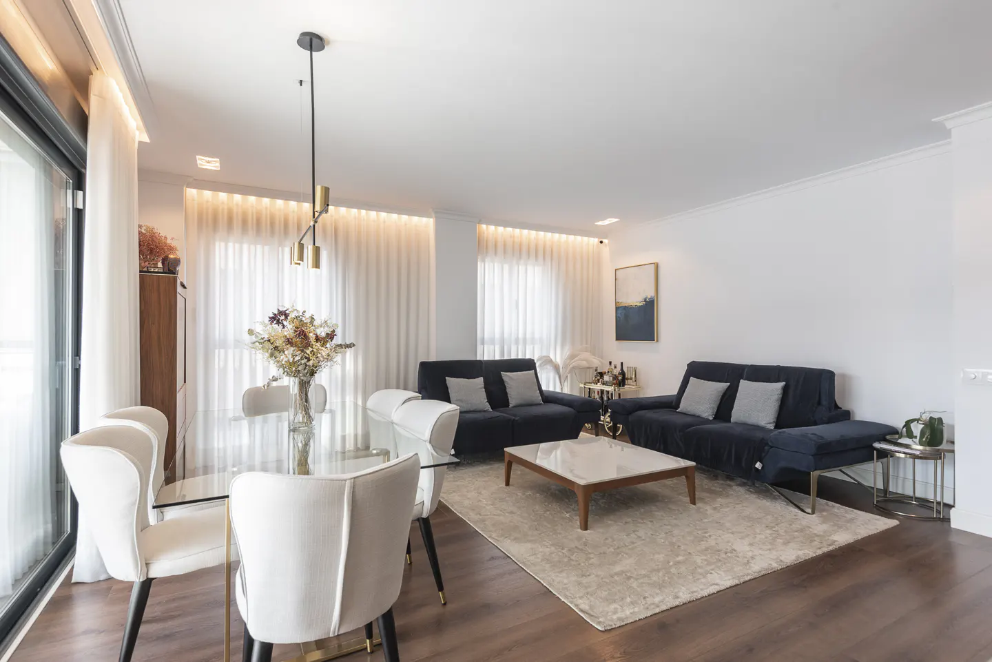 Bright living room with a glass table, white chairs, two blue sofas, and a light beige rug on a dark wood floor.
