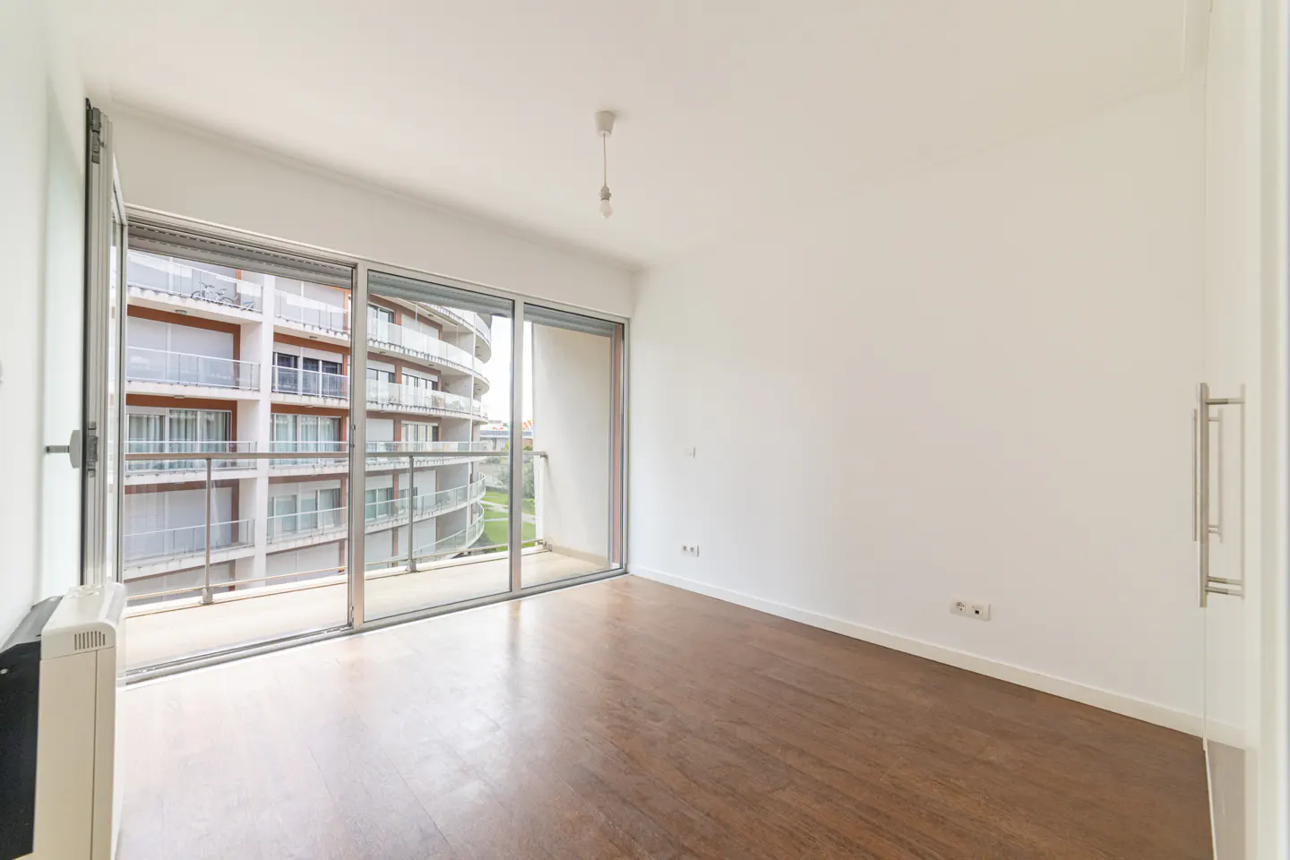 Bright, empty room with wood floors and white walls. Large sliding glass doors open to a balcony with a view of a modern apartment building.