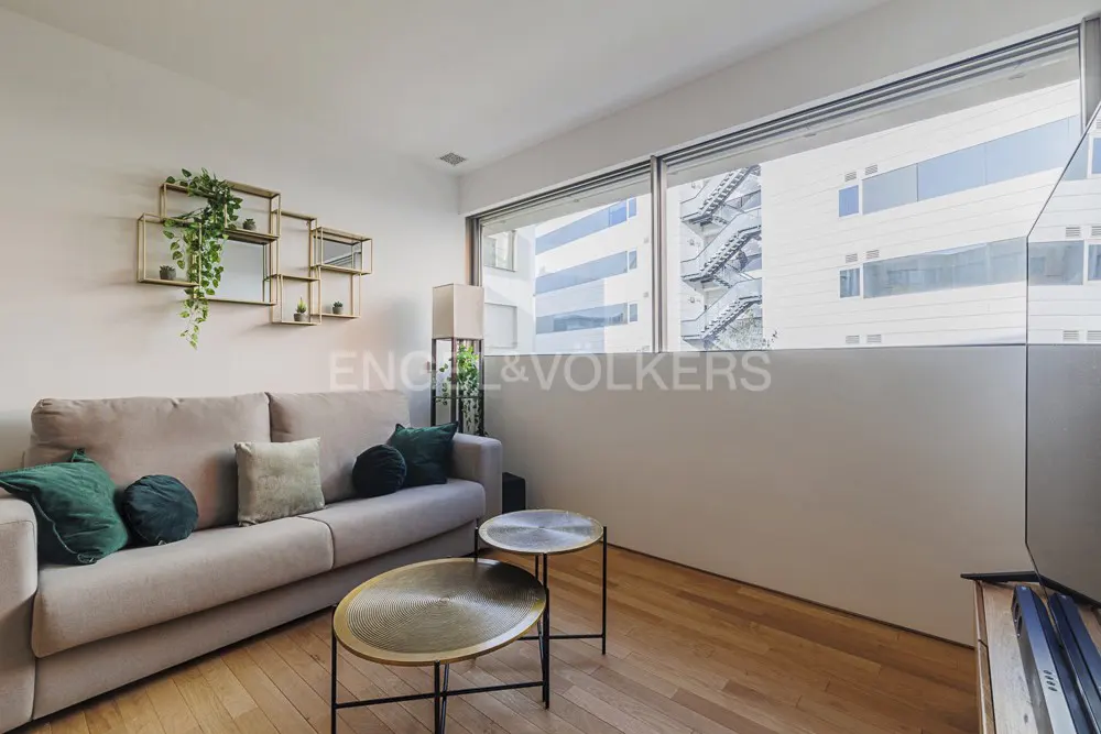 Living room with a beige sofa, green pillows, two round tables, and a gold geometric shelf with plants. Large windows show a building exterior.