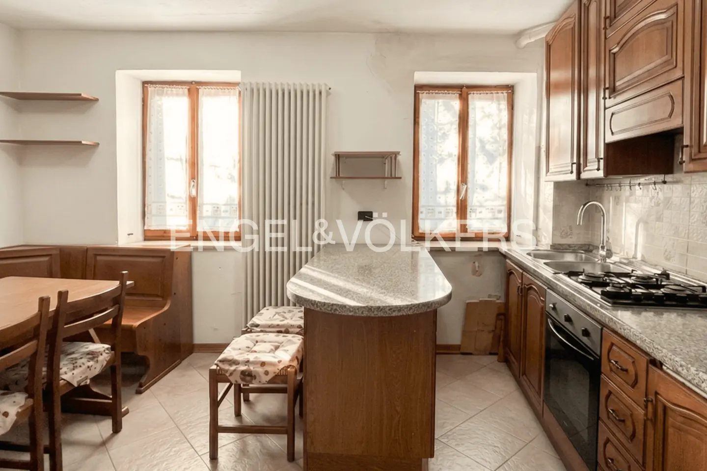 Kitchen with wooden cabinets, granite island, dining nook with bench seating, and two windows with white curtains.