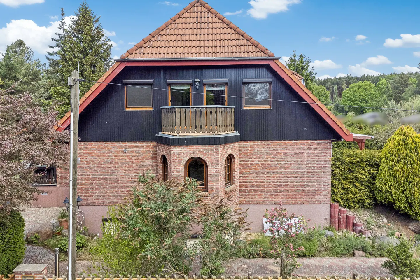Two-story brick house with a balcony, black wood siding, and a red tile roof, surrounded by trees.