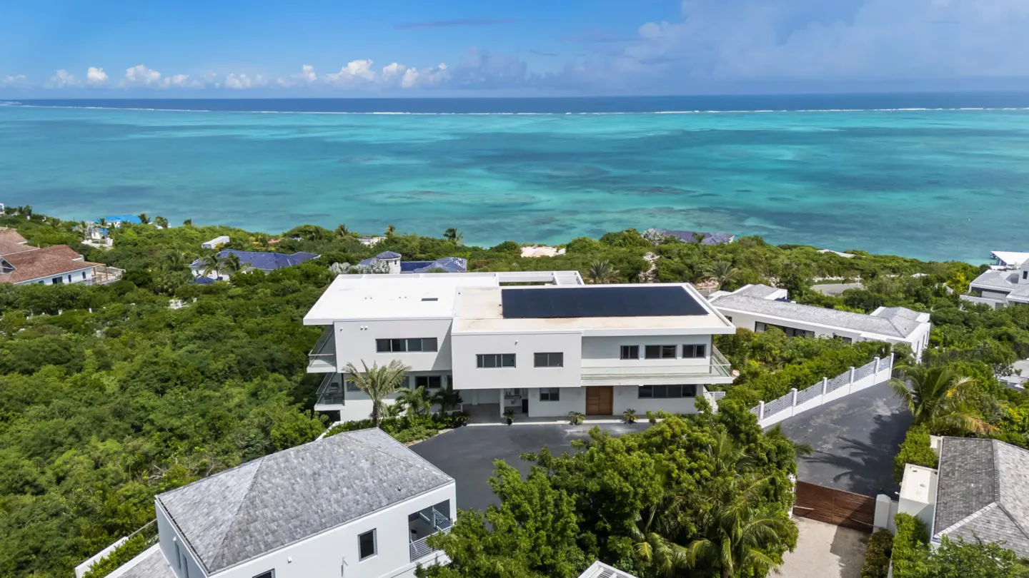 Aerial view of a modern white house with solar panels, surrounded by lush green trees and turquoise ocean.