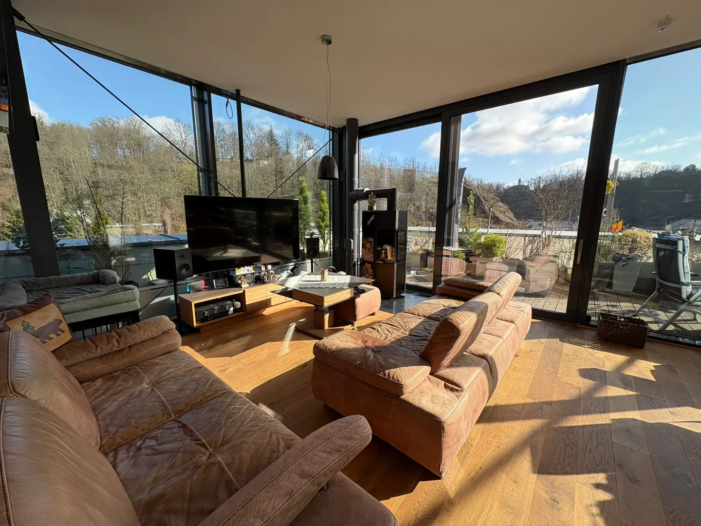 Bright living room with brown leather sofas, wood floors, and floor-to-ceiling windows overlooking trees and a blue sky.