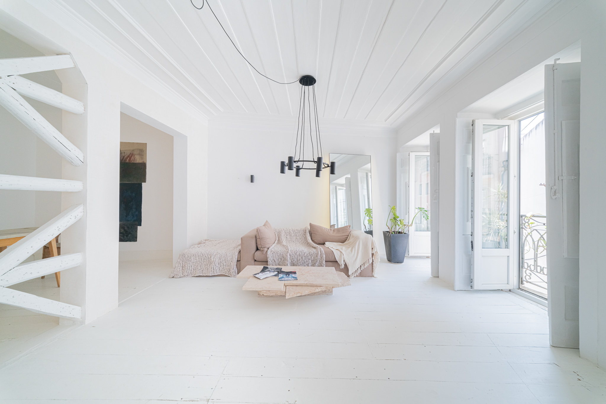 Bright, minimalist living room with white walls, wooden floors, a beige sofa, a modern black chandelier, and balcony doors.