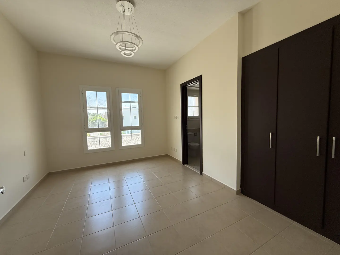 A bright, empty room with beige walls, tile floors, and a modern chandelier. Two windows let in natural light. A dark wood closet and bathroom door are on the right.