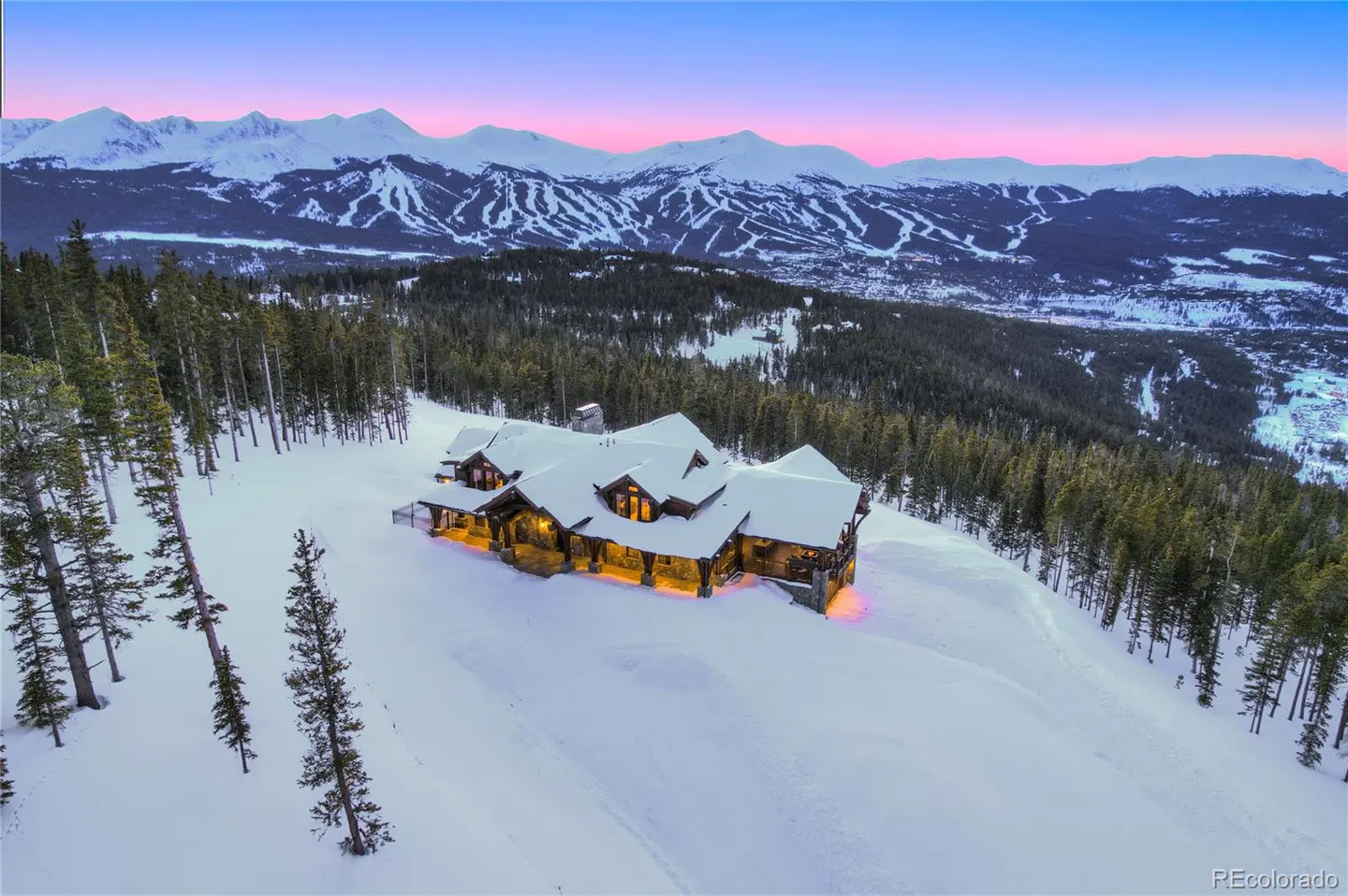 A large, snow-covered log home sits on a hill, surrounded by pine trees. Mountains are in the background under a pink and blue sky.