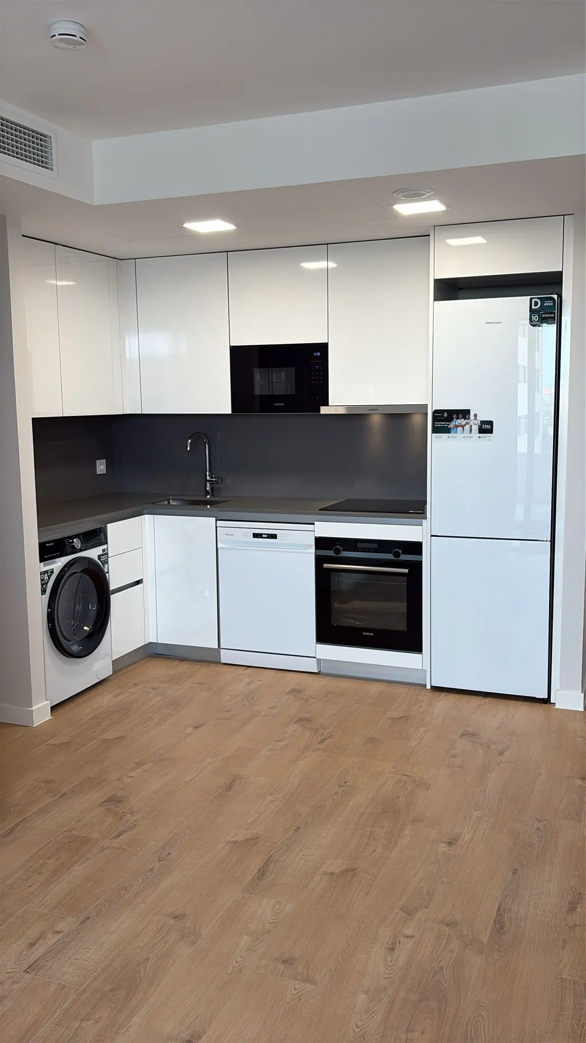Modern kitchen with white cabinets, black countertops, and wood floors. Appliances include a washing machine, dishwasher, oven, microwave, and refrigerator.