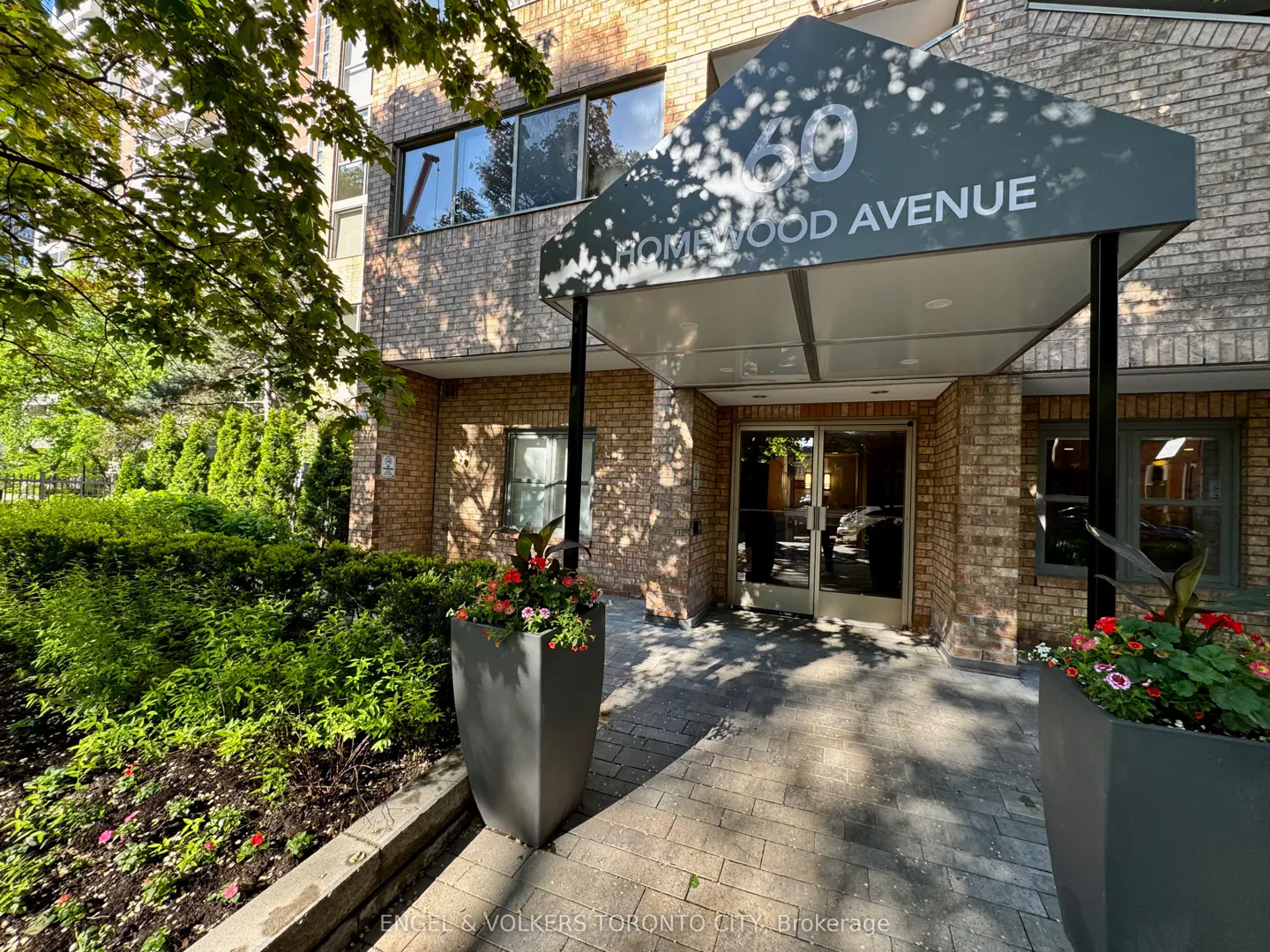 Entrance to 60 Homewood Avenue with a dark awning, brick facade, glass doors, and large planters with colorful flowers.