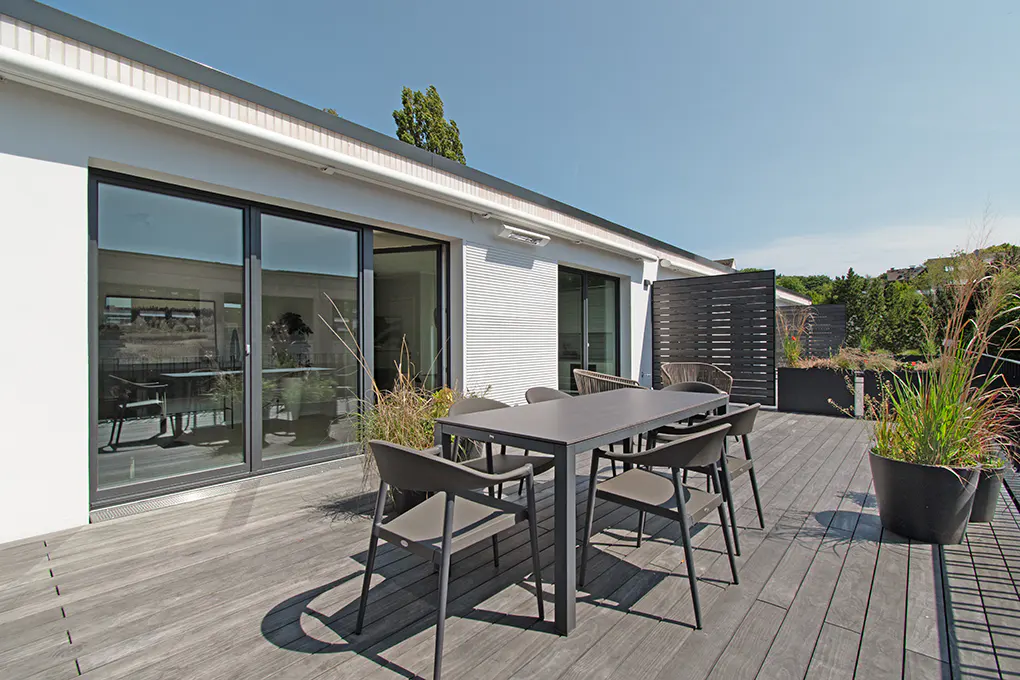 Outdoor patio with a gray table and chairs on a wooden deck. Sliding glass doors lead into a white building. Plants in pots add greenery.