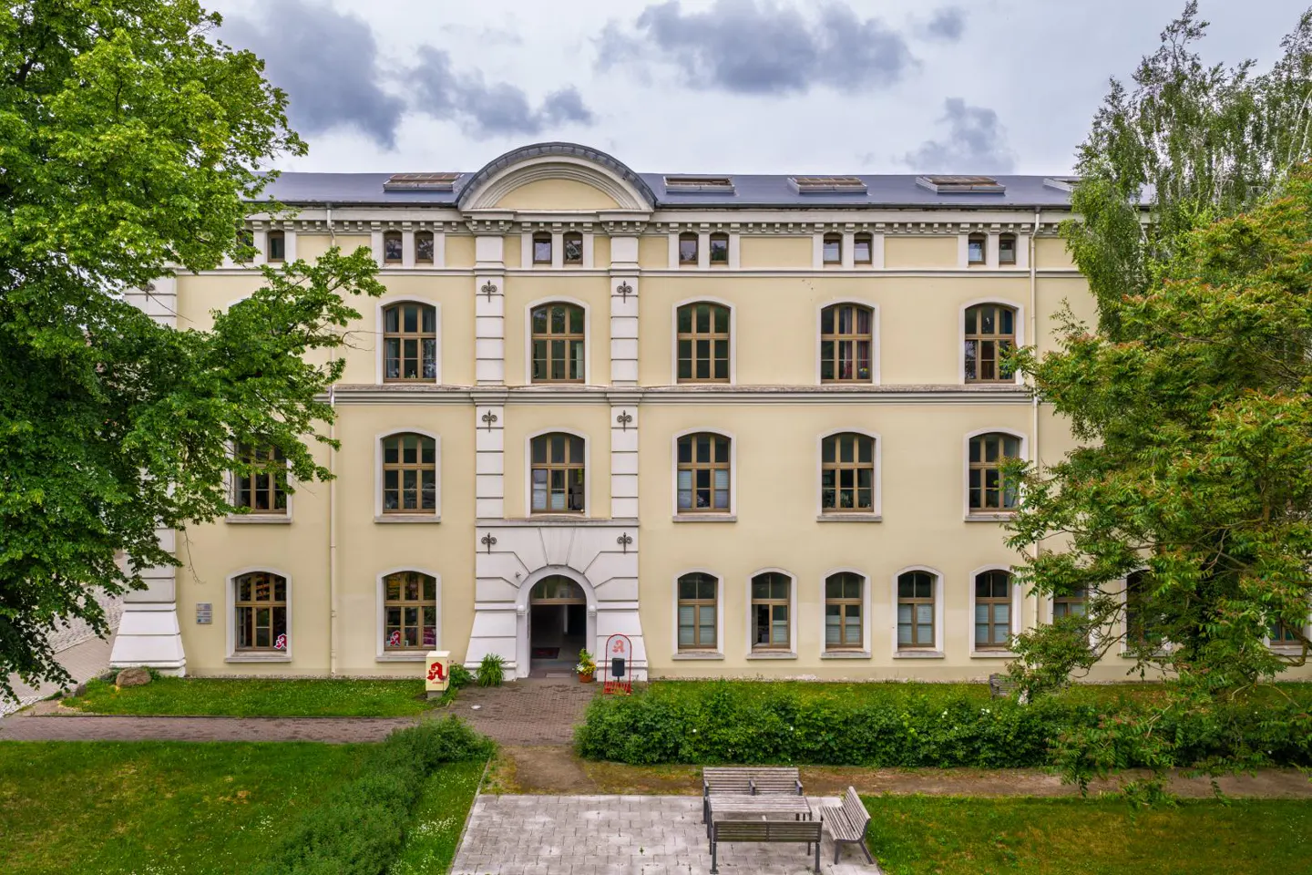 A three-story, cream-colored historic building with arched windows, surrounded by green trees and a small park area.