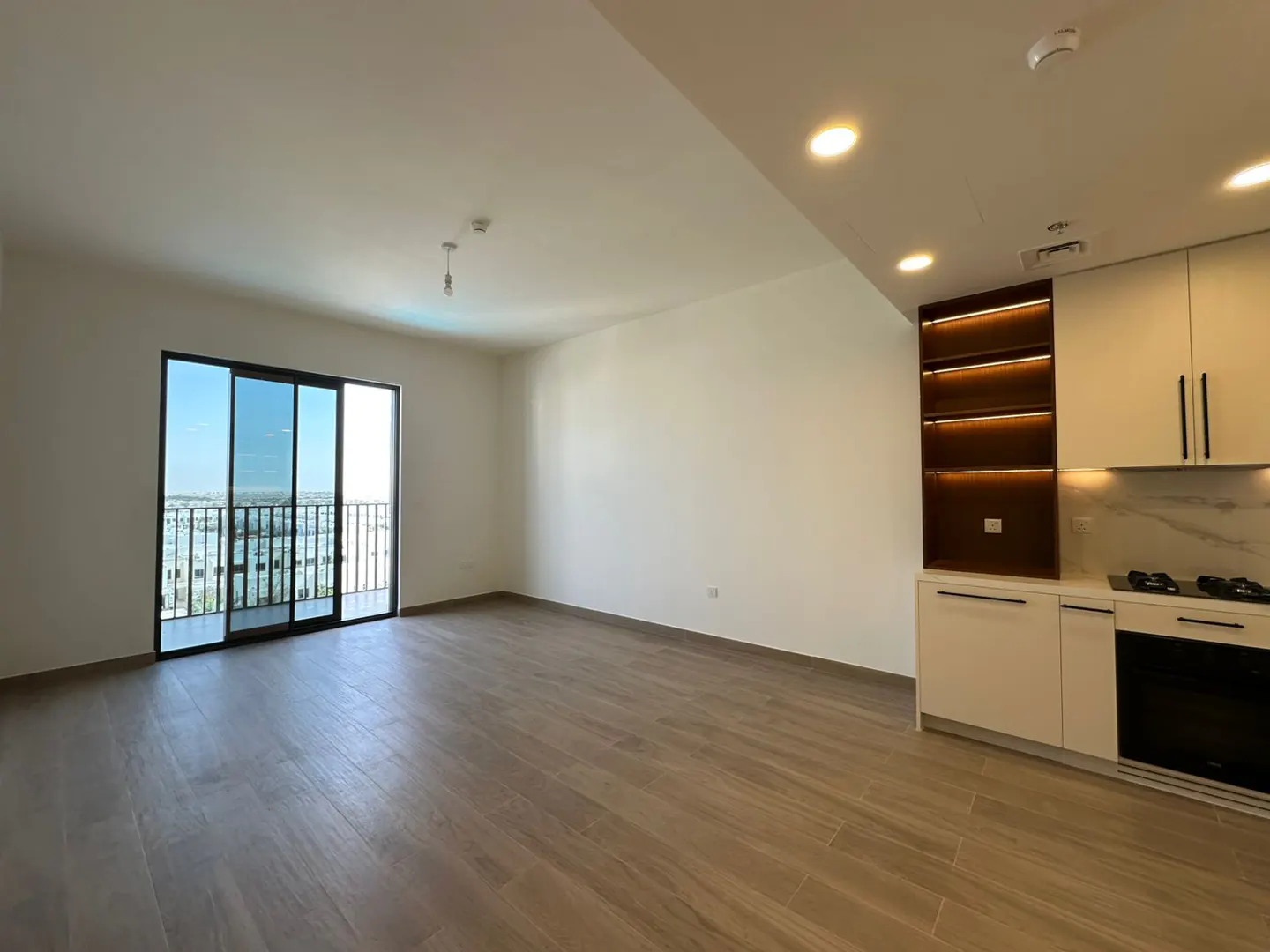 An empty apartment with wood floors, white walls, and a sliding glass door to a balcony. A modern kitchen with white cabinets is on the right.