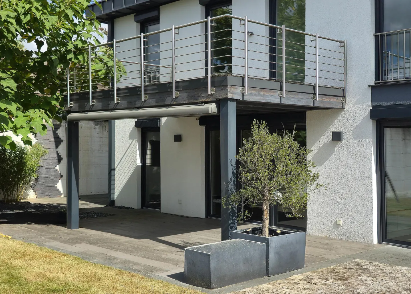 Modern home exterior with a balcony, stainless steel railings, and gray stone patio. A small tree sits in a square planter.