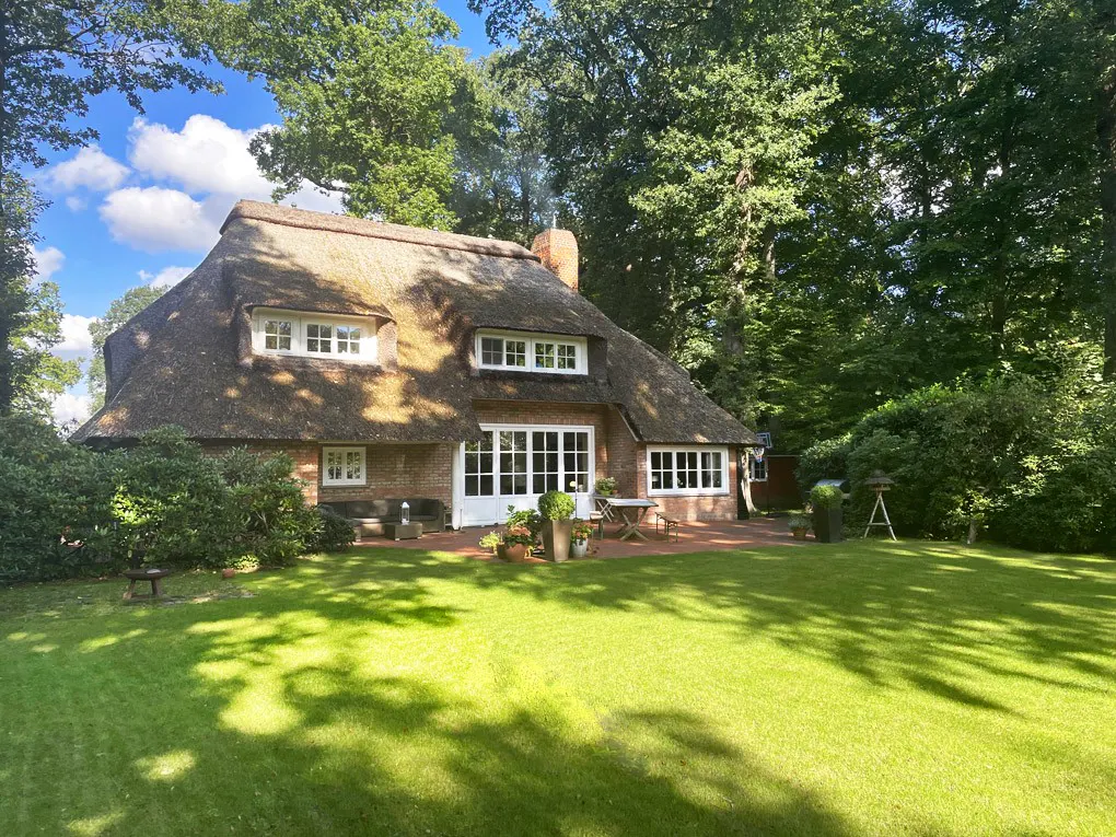 Exterior view of a brick house with a thatched roof, white trim, and a green lawn surrounded by trees.