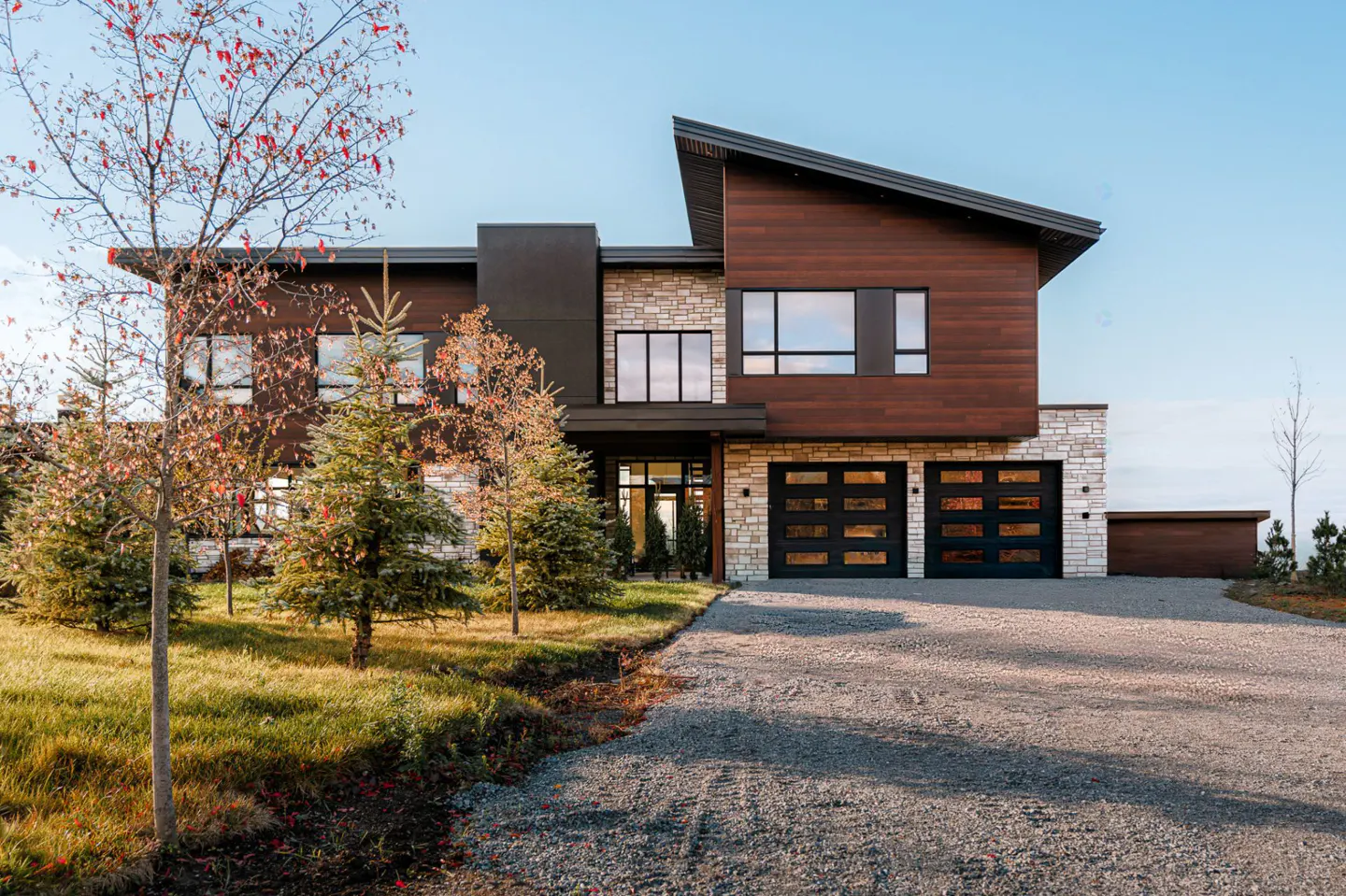 Modern two-story home with wood and stone facade, black garage doors, and gravel driveway. Trees in the front yard.