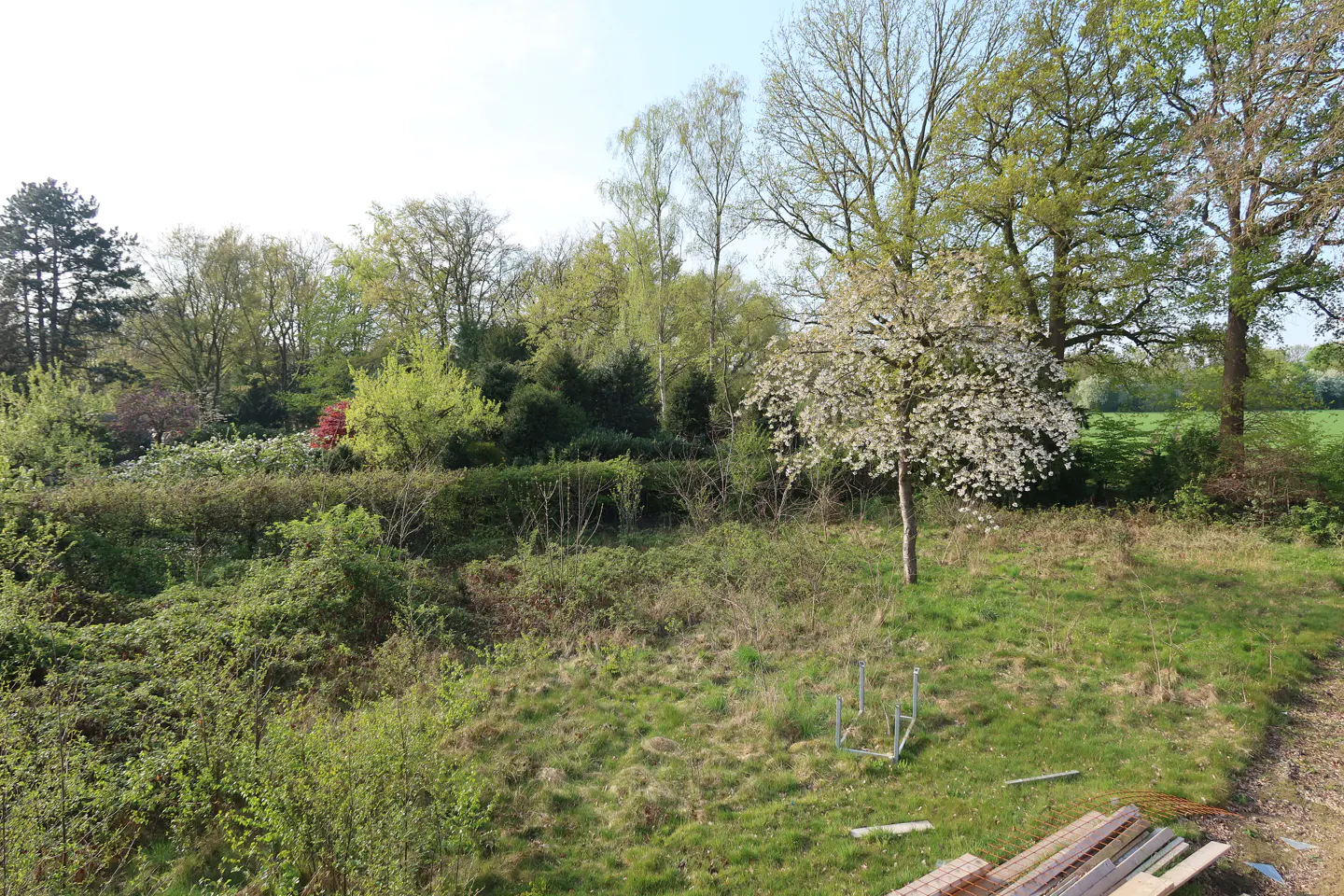 A lush green landscape with trees, bushes, and a flowering tree in the foreground. Construction materials are visible at the bottom.
