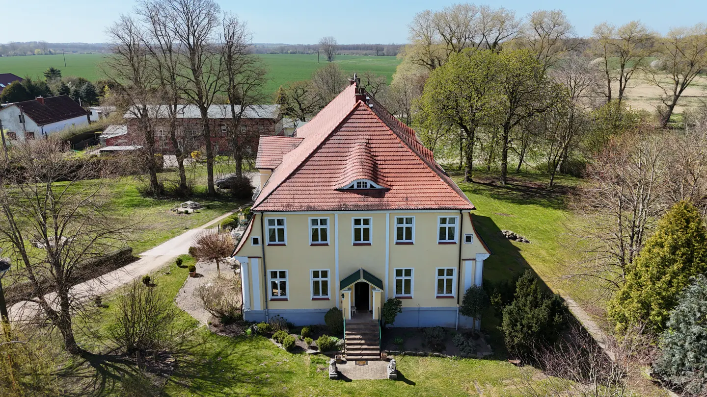 Aerial view of a yellow two-story house with a red tile roof, surrounded by green grass and trees.