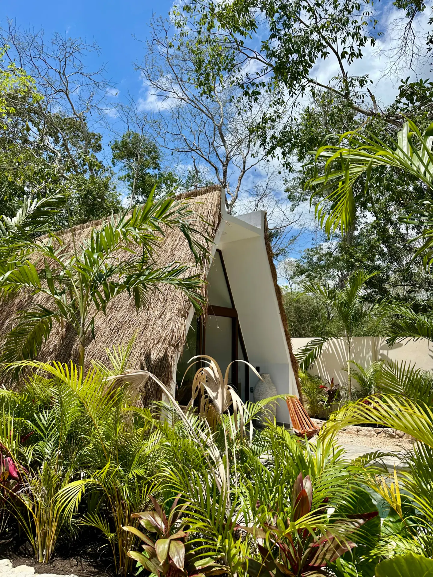A-frame cabin with a thatched roof, surrounded by lush green tropical plants under a blue sky.