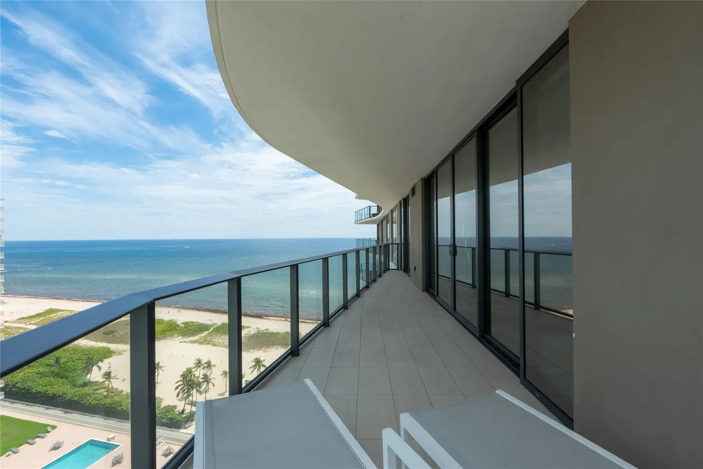 Condo balcony with ocean view. Glass railings, lounge chairs, and sliding glass doors. Blue sky and water.