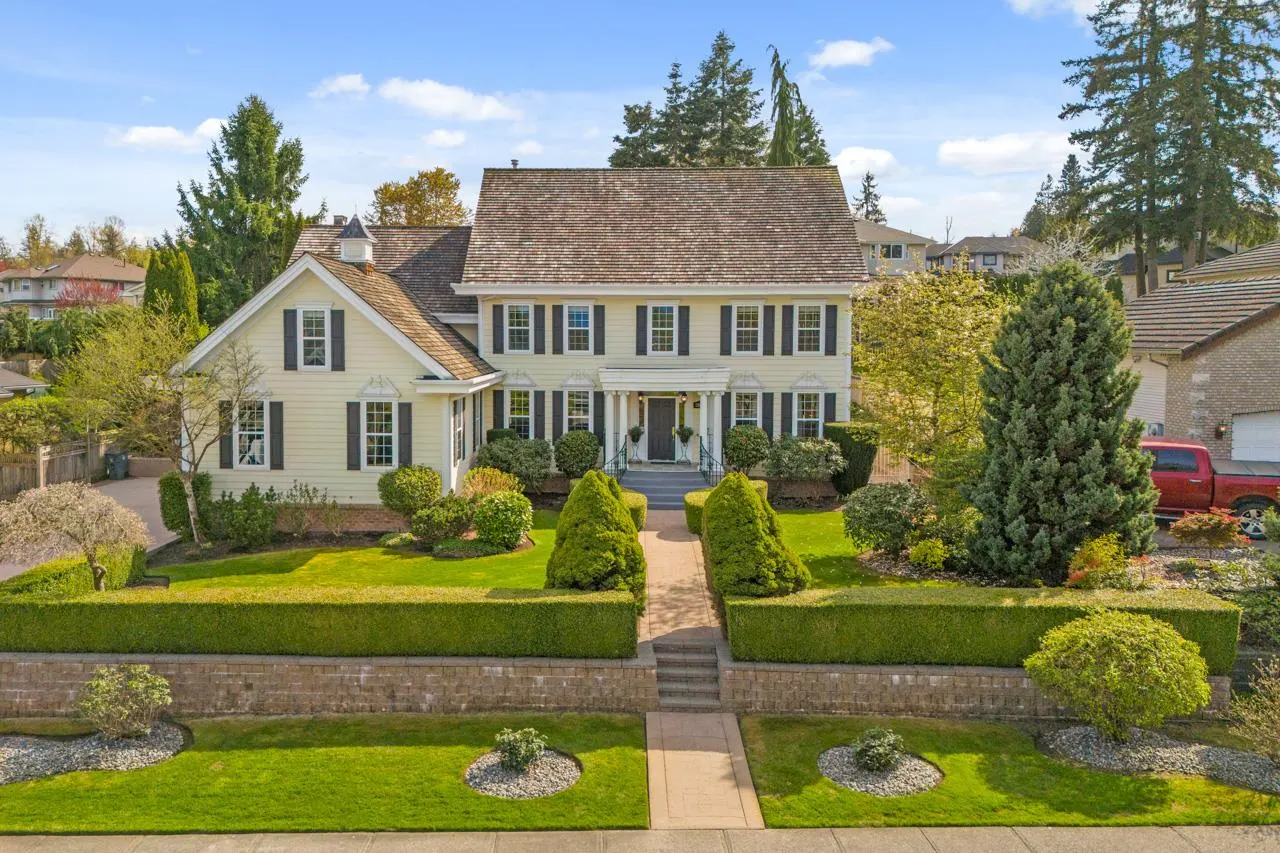 Two-story yellow house with black shutters, a brown roof, and manicured green landscaping.