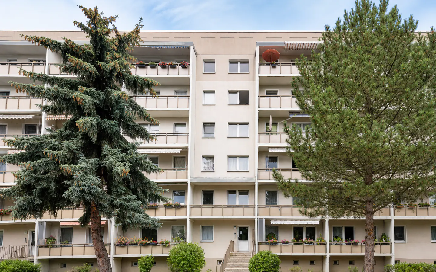 A beige apartment building with balconies and two large green trees in front.