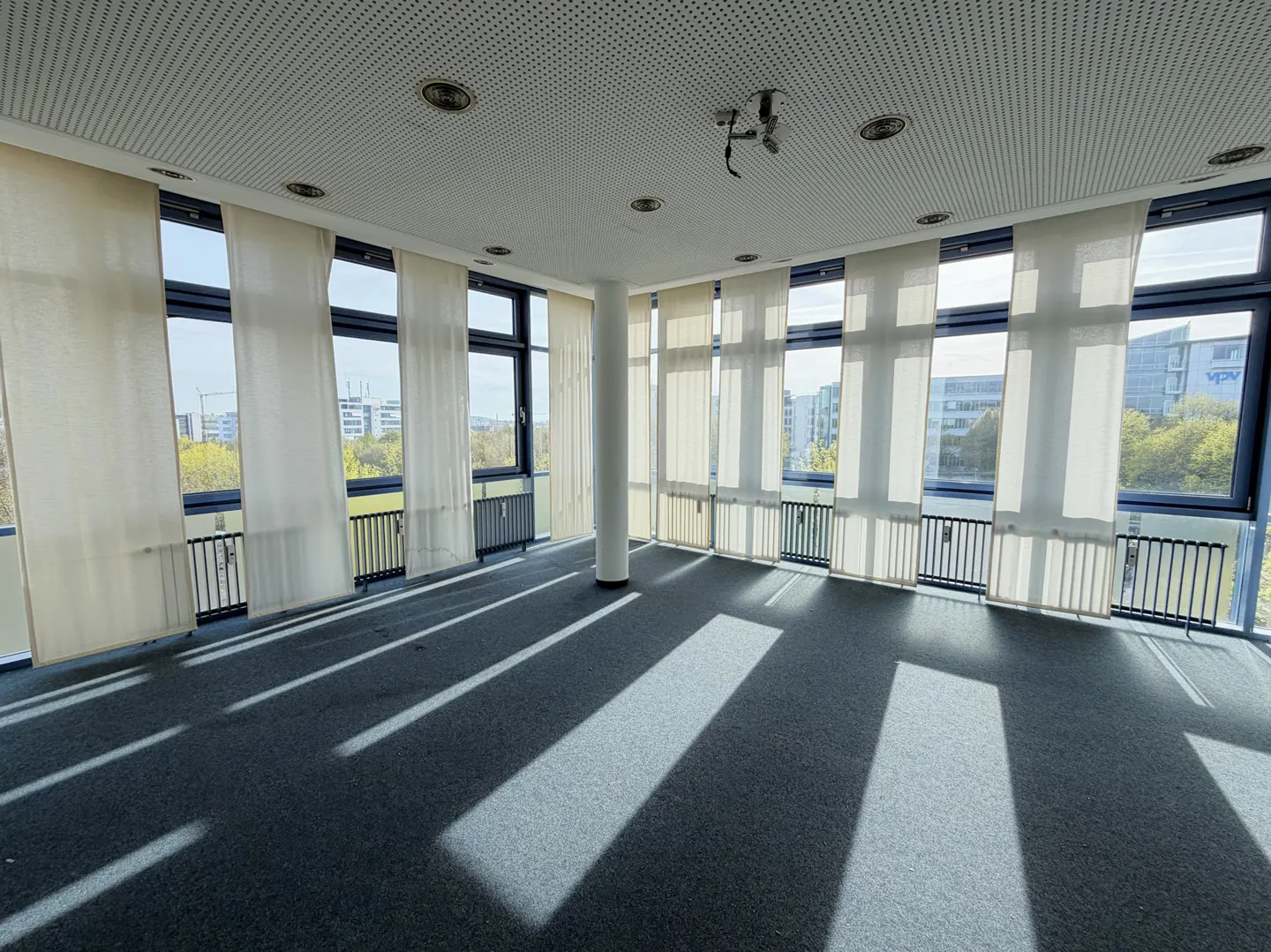 Empty office space with gray carpet, white curtains, and large windows. Sunlight streams in, creating shadows on the floor.