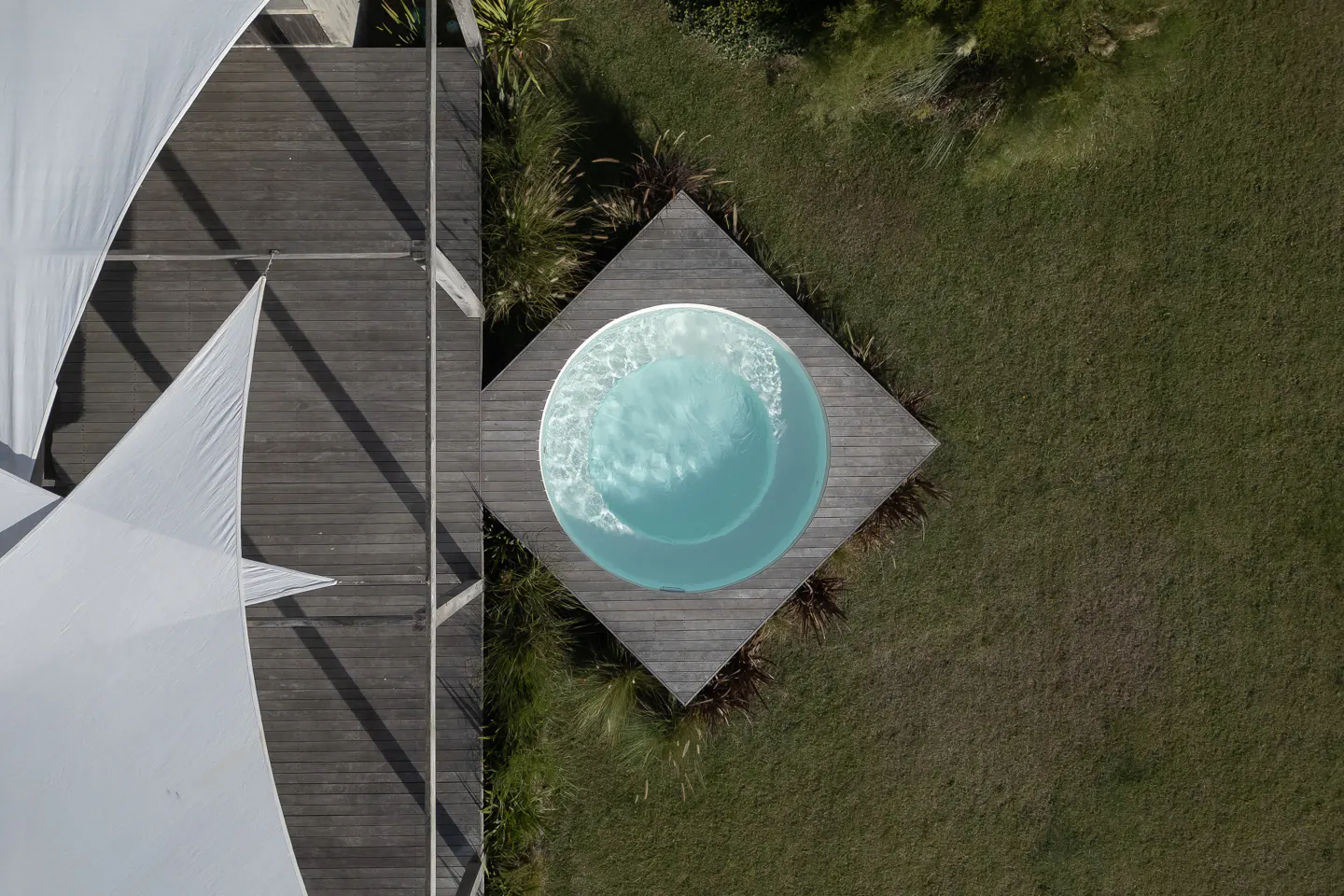 Aerial view of a round, light blue pool on a square wooden deck, next to a wooden deck with white sunshades and green grass.
