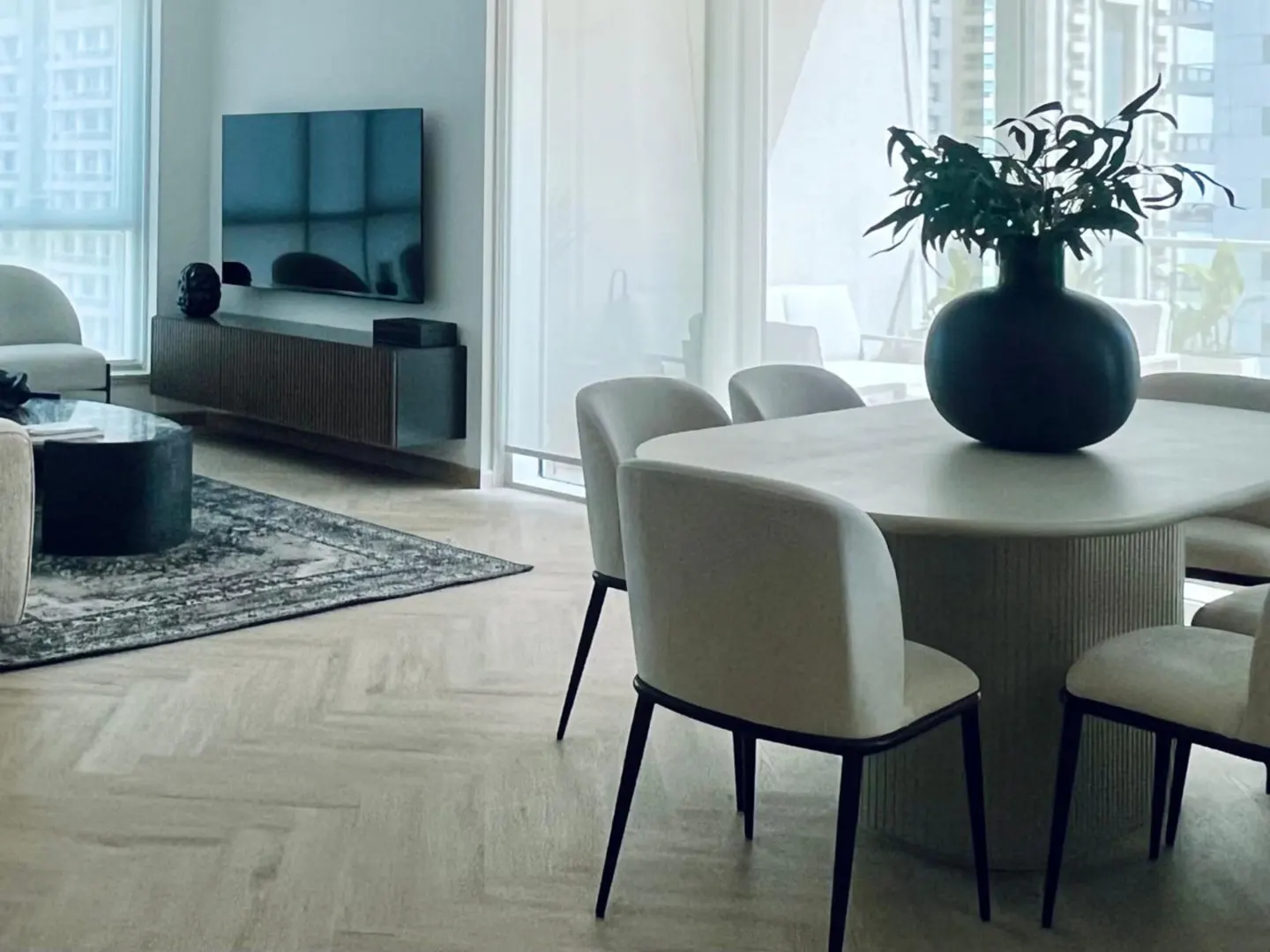 A modern apartment interior with a dining table, chairs, and living room area. A black vase with greenery sits on the table. A TV is mounted on the wall.