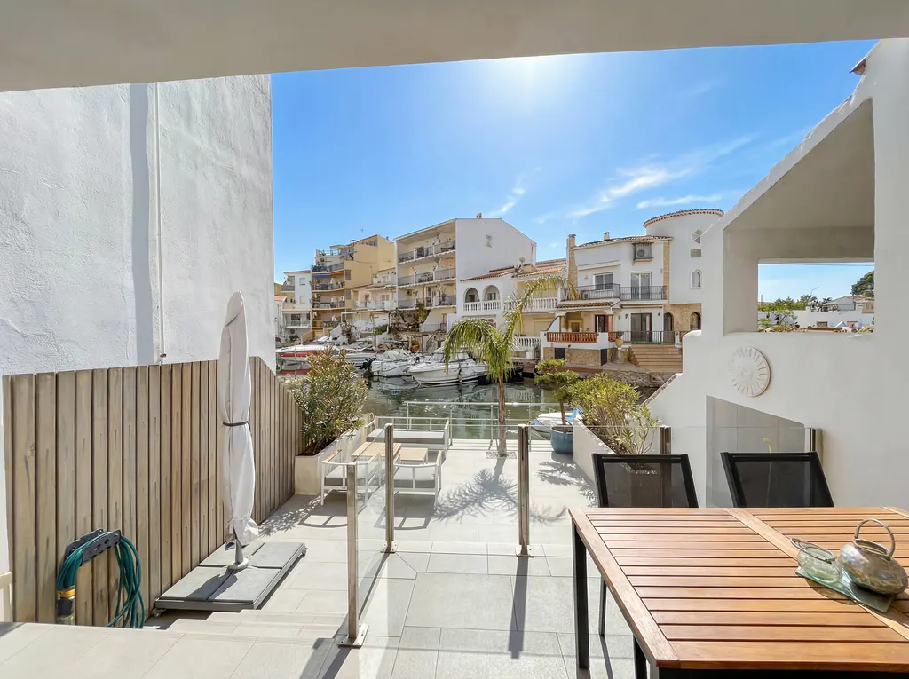 View from a patio with a wooden table and chairs overlooking a canal with boats and white buildings under a blue sky.