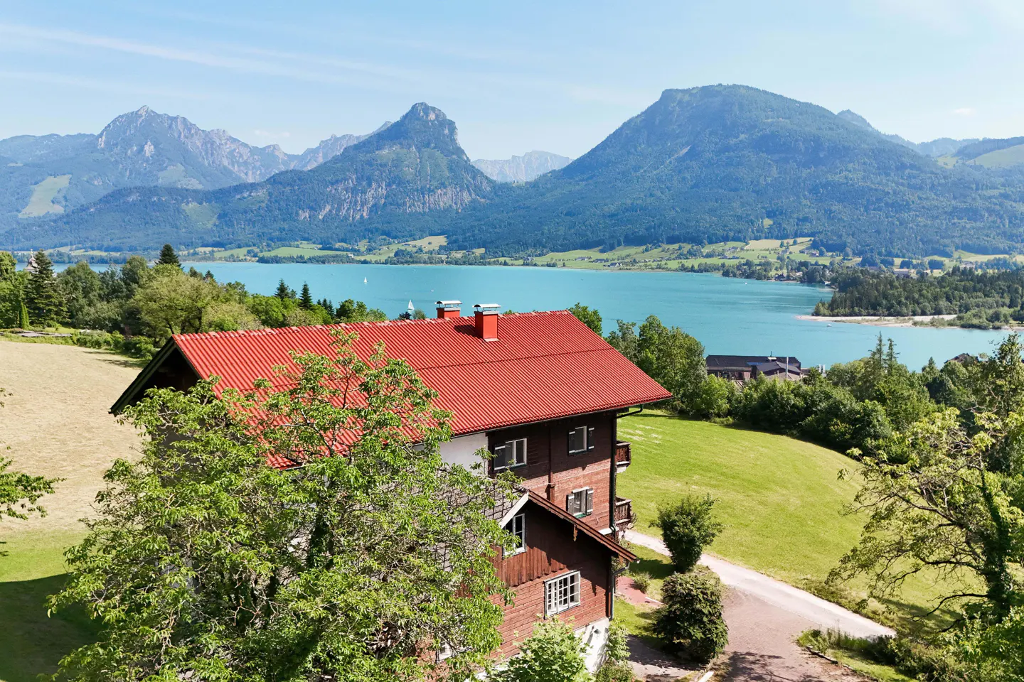 Scenic view of a brown house with a red roof, green lawn, turquoise lake, and mountains in the background.